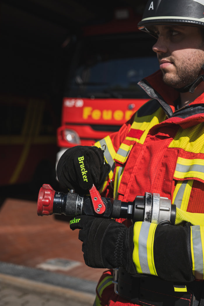 Feuerwehrmann hält mit schwarzen Feuerwehrhandschuhen S Black aus Leder und Kevlar, mit gelb/grau Reflektoren und gelben Brucker Stick auf dem Zeigefinger ein Strahlrohr mit Schlauch vor einem Feuerwehreinsatzfahrzeug