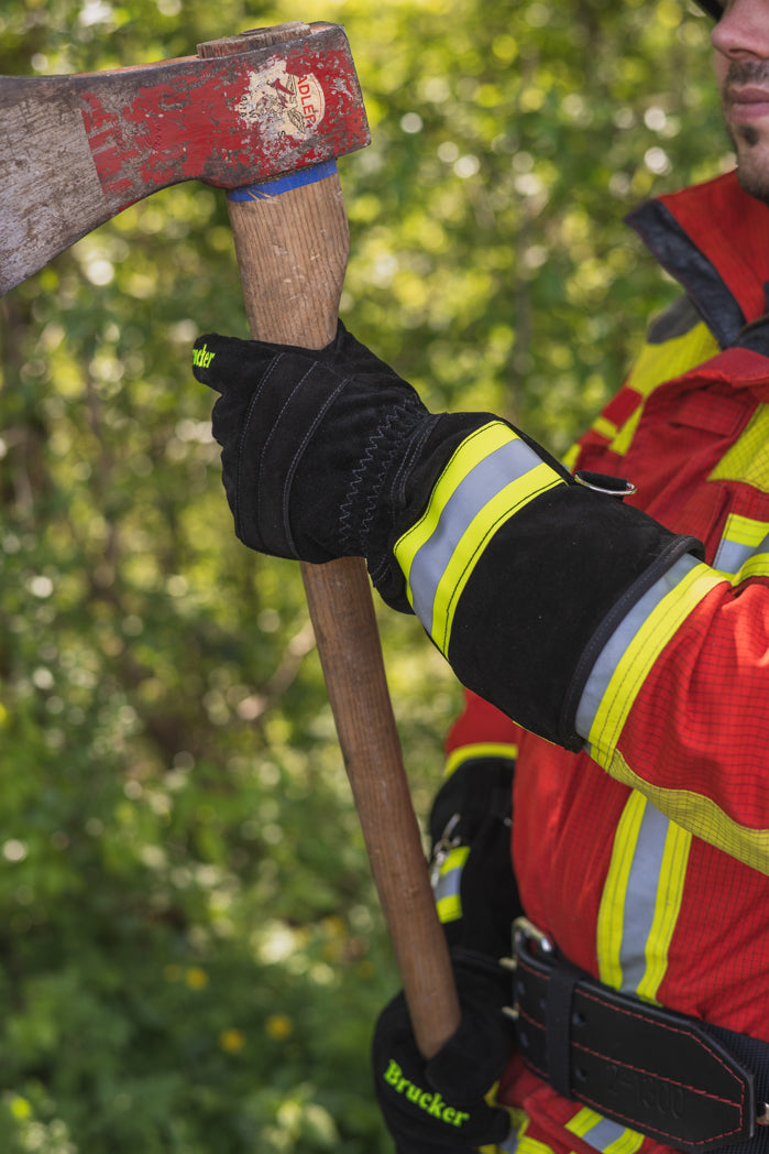 Feuerwehrmann hält mit Feuerwehrhandschuhen S Black eine Axt vor einem Gebüsch
