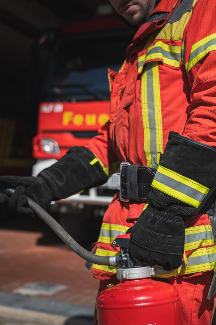 Feuerwehrmann hält mit schwarzen Feuerwehrhandschuhen S Black aus Leder und Kevlar, mit langer Stulpe mit gelb/grau Reflektoren und gelben Brucker Stick auf dem Zeigefinger einen Feuerlöscher vor einem Feuerwehreinsatzfahrzeug