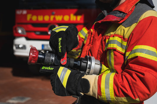 Feuerwehrmann hält mit schwarzen Feuerwehrhandschuhen Black aus Leder und Kevlar, mit gelb/grau Reflektoren und gelben Brucker Stick auf dem Zeigefinger ein Strahlrohr mit Schlauch vor einem Feuerwehreinsatzfahrzeug